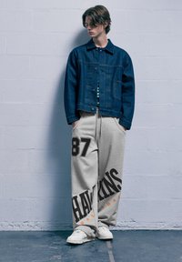 Farmerdzseki gombos záródással, szürke melegítőnadrág nagy „87”-es számmal és „HAWKINS BASKETBALL” felirattal fekete és narancssárga színben, fehér cipőkkel párosítva.