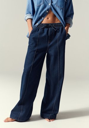 Pantalones de mezclilla anchos en un tono azul oscuro con cintura elástica, detalle de cordón y costuras en contraste. Combinados con una camisa de mezclilla de botones.