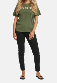 Woman wearing an olive green T-shirt with "HOPE" text, black slim-fit pants, and black flip-flops standing against a white background.