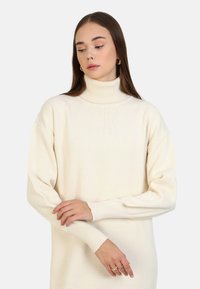 Jersey de cuello alto de color beige claro con un diseño holgado, mangas anchas y acentos de canalé en el cuello y los puños, confeccionado en una tela texturizada.