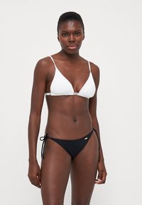 White triangle bikini top with thin straps, paired with a black ribbed bikini bottom featuring ties. Smooth fabric and minimalist design.