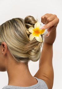 Hårklämma i form av en gul och vit blomma, tillverkad av glänsande plast, håller upp en knut med ljust brunt hår mot en neutral bakgrund.