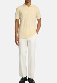 Polo in cotone giallo pallido con colletto classico e maniche corte, abbinato a pantaloni color crema e calzature nere.