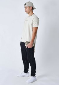 Camisa beige de manga corta con detalle de cadena, pantalones cargo negros y zapatillas blancas. Sosteniendo un bolso con un patrón texturizado y una correa.