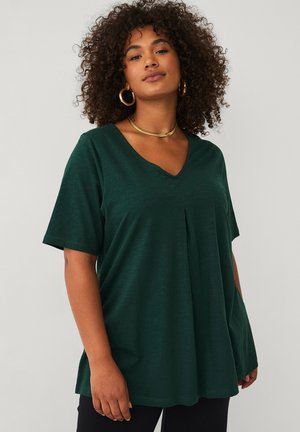 Kvinde med krøllet hår iført guldringeøreringe og en halskæde, iført en mørkegrøn T-shirt med korte ærmer og V-udskæring foran med plissering.