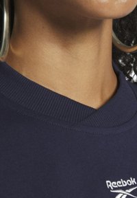 Námořnický modrý svetr s žebrovaným kulatým výstřihem, vyšívaným logem Reebok a měkkou texturou látky. Detailní pohled na límec a oblast krku.