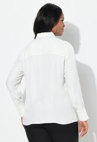 Blusa bianca a maniche lunghe con colletto, texture liscia, polsini con bottoni e orlo curvo, vista dal retro.