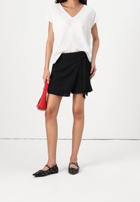 Kobieta w białej bluzce z krótkim rękawem i dekoltem w serek, czarnych szortach z zakładką, czarnych balerinach Mary Jane z ćwiekami, trzymająca małą czerwoną torebkę przy boku.