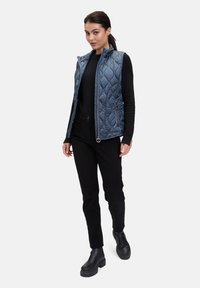 Gequiltede, lichtblauwe vest met hoge kraag, met ritszakken, gedragen over een zwarte top en zwarte broek; model draagt zwarte laarzen.
