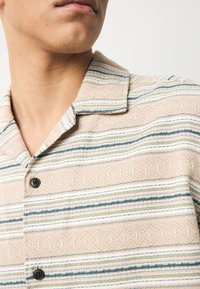 Camicia a maniche corte realizzata in tessuto strutturato, con un motivo geometrico a righe beige, blu e verdi e bottoni neri sulla patta.