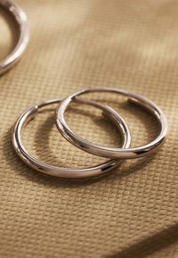 Zilveren hoop oorbellen met een gladde, glanzende oppervlakte, met een eenvoudig cirkelontwerp. Geplaatst tegen een gestructureerde beige achtergrond.