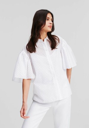 Camicia bianca con maniche corte a sbuffo, caratterizzata da un motivo testurizzato. Realizzata in tessuto leggero, abbinata a pantaloni bianchi.