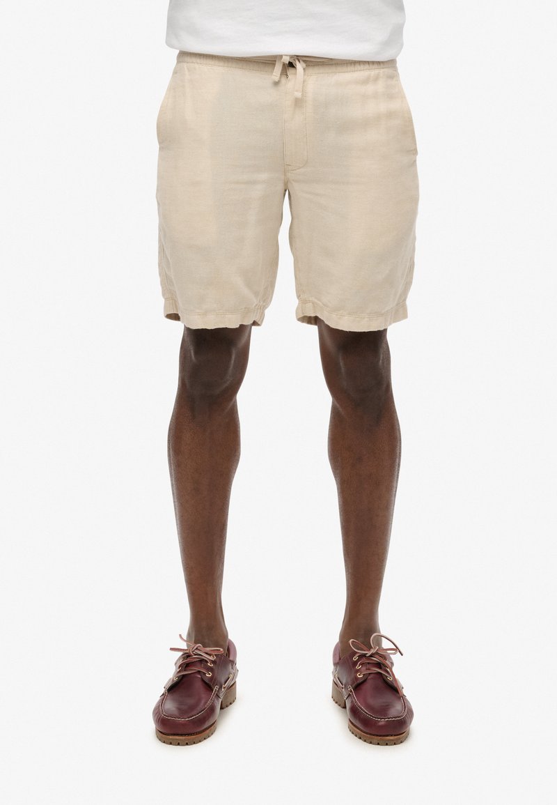 Pantalones cortos de lino beige con cintura elástica y cordón, que presentan un ajuste relajado y puños doblados, combinados con zapatos burdeos.