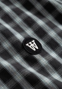 Tissu à carreaux noirs et gris avec un patch rond noir présentant en son centre les lettres stylisées "AW" brodées en blanc.
