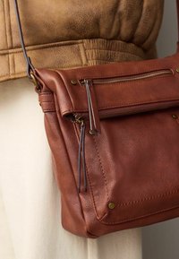 Smeđa kožna torba za rame sa zippered prednjim džepom, brass hardverskim detaljima, šivenim ukrasima i ravnom bazom za stabilnost.