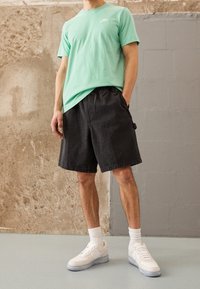 Třpytivě zelené bavlněné tričko, černé džínové šortky a bílé tenisky s průhlednou podrážkou. Volný střih a uvolněný design.