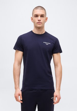 Fiatal férfi rövid hajjal, sötétkék Björn Borg pólót és hozzáillő nadrágot viselve, egy egyszerű világos háttér előtt áll.