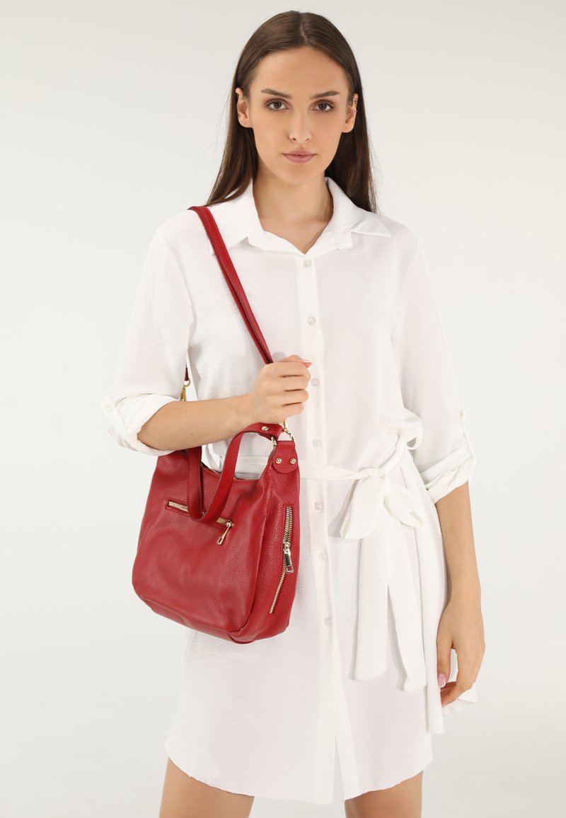 Rote Lederhandtasche mit strukturierter Form, ausgestattet mit zwei Reißverschlusstaschen und einem Schulterriemen, gehalten von einer Person in einem weißen Hemdkleid.