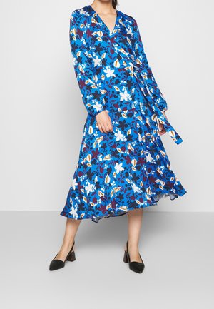 Vrouw die een blauwe midi wikkeljurk draagt met witte, zwarte en gele bloemmotieven en lange mouwen, gecombineerd met zwarte schoenen met lage hak.