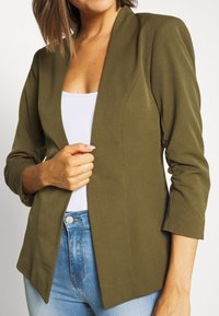 Olive green blazer with a tailored fit, long sleeves, and a notch collar. Fabric appears smooth and structured, paired with a white top and blue jeans.