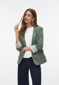Groene blazer met een getailleerde pasvorm, voorzien van gestreepte manchetten, gedragen over een witte top en gecombineerd met donkere spijkerbroek. Glad stoftextuur.