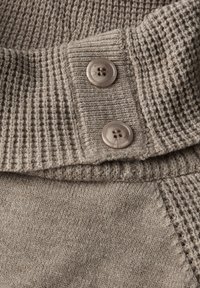 Gebreide trui in lichtbruin met een gestructureerde, geribbelde kraag. Beschikt over twee bruine knopen en een gladde stof met subtiele stikseldetails.