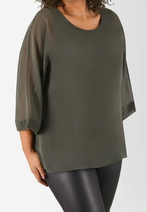 Vrouw draagt een losse donkergroene blouse met driekwart doorzichtige mouwen en zwarte nauwsluitende broek met leren uitstraling tegen een effen achtergrond.