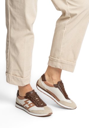 Osoba nosząca beżowe spodnie sztruksowe oraz brązowo-beżowo-białe sneakersy na sznurowadła z wzorem "49" po bokach.