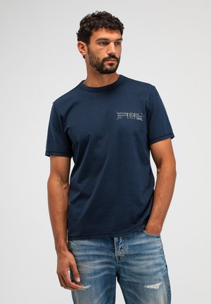 Man met baard die een marineblauw grafisch T-shirt en versleten blauwe spijkerbroek draagt, staand met één hand in de zak.
