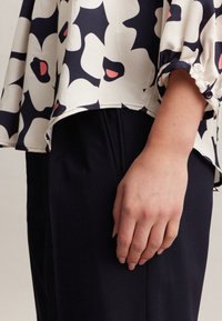 Blusa leggera con un motivo floreale in crema, blu navy e rosa, abbinata a pantaloni scuri. Presenta un orlo a goccia e maniche a risvolto.