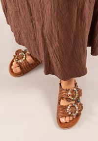 Sandalias trenzadas marrones con adornos decorativos, que presentan grandes acentos circulares y tres correas ajustables. Suela texturizada y plana.