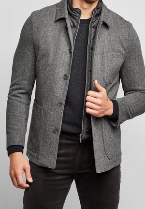 Homme portant une veste en laine gris à chevrons superposée à une veste noire matelassée zippée et un pull noir, ajustant la veste extérieure avec la main droite.