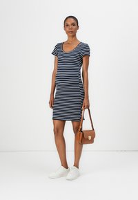 Vestido listrado azul-marinho com decote redondo, mangas curtas e silhueta ajustada. Ténis brancos e uma pequena bolsa castanha completam o visual.