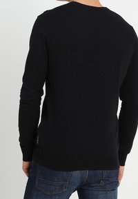 Zwarte gebreide trui met lange mouwen, ronde halslijn en geribbelde zoom. Draagt over donkerblauwe jeans. Heeft een stoffen label aan de zijkant.