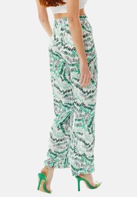 Flowy, high-waisted trousers in white with green and black abstract print. Features a smooth texture and a wide-leg design. Worn with green heels.