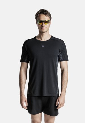 Uomo che indossa una maglietta sportiva nera, pantaloncini neri e occhiali da sole sportivi riflettenti, in piedi davanti a uno sfondo bianco.