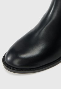 Primo piano di una punta di stivale in pelle nera con texture a grana fine e suola a welt cucita su uno sfondo chiaro.