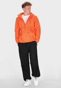 Giacca arancione con motivo astratto nero, colletto con zip, abbinata a pantaloni neri e scarpe da ginnastica bianche. Design solido, look moderno e casual.
