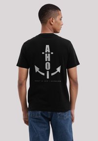 Black cotton t-shirt featuring a silver graphic on the back with the text "AHOI" and an anchor design, accented by arrows and brand name.