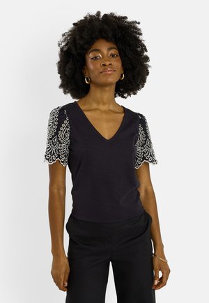 Mujer con peinado afro que lleva una camiseta negra con escote en V, mangas cortas de encaje blanco y pantalones negros, de pie frente a un fondo sencillo.