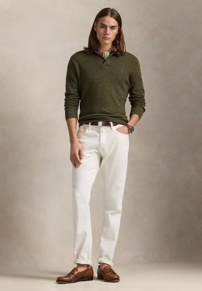 Nuori mies olkapäähän ulottuvilla hiuksilla, yllään oliivinvihreä neulepaita, värikäs kaulahuivi, valkoiset housut, ruskea vyö, ruskeat loaferit ja rannekello.