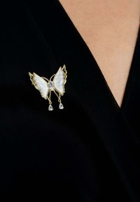 Gold butterfly brooch with white enamel wings, a central clear gem, and two hanging clear teardrop accents on a dark fabric background.