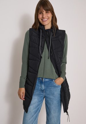 Femme souriante debout, portant un gilet matelassé noir sans manches sur un pull vert zippé et un jean bleu clair, une main dans la poche.