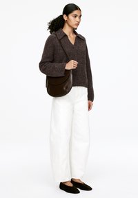 Frau mit lockigem Haar, die einen dunkelbraunen Pullover, weiße weite Hosen, schwarze Ballerinas trägt und eine dunkelbraune Leder-Umhängetasche dabei hat.