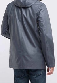 Man i grå huvtröja av vattentåligt material och blå jeans, sedd bakifrån mot en enfärgad bakgrund.