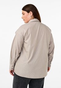 Ylpeä raidallinen oversized napitettava paita vaaleanbeige värissä, jossa on pystysuorat viivat, klassinen kaulus ja napitettu hihansuu. Sileä rakenne.