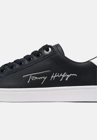 Baskets basses noires avec semelle blanche, lacets noirs, œillets argentés et signature métallique "Tommy Hilfiger" sur le côté.