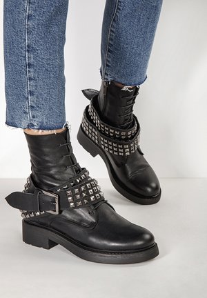 Czarne skórzane botki za kostkę posiadają dekoracyjne srebrne ćwieki oraz mocną klamrę, niską obcas i detale sznurowania. Noszone z postrzępionymi dżinsami.
