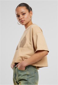 Beiges lõhustatud T-särk, mis on valmistatud puuvillast, koos pruuni graafilise kujundusega. Kombineeritud teksapükstega, mis rõhutavad taskuid ja millel on mugav lõige.