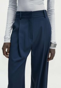 Pantalon bleu marine à coupe ajustée, avec plis, texture lisse et taille haute, associé à un haut ajusté à manches longues.